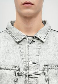 Νέος άτομο φοράει ένα ανοιχτό γκρι τζάκετ ντένιμ με οξέα πλύση, με γιακά και ασημένια κουμπιά, που φαίνονται λεπτομέρειες στο λαιμό και το κάτω μέρος του προσώπου.