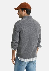 Grijze wollen trui met een hoge kraag, gedragen over een blauw-wit gestreept shirt. Bruine corduroy pet. Zichtbare spijkerbroek.