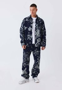 Hombre vistiendo una chaqueta y pantalones de patrón pixelado en azul oscuro y gris, camiseta gráfica blanca y zapatillas blancas, de pie frente a un fondo blanco.