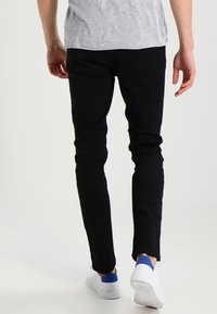 Personne portant un pantalon noir, un t-shirt gris et des baskets blanches avec des accents bleus, marchant loin sur un fond clair uni.