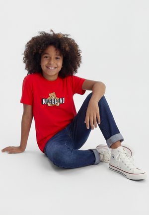 Enfant aux cheveux bouclés assis par terre en souriant, portant un t-shirt rouge avec un logo d'ours en peluche, un jean bleu retroussé aux chevilles et des baskets blanches montantes.