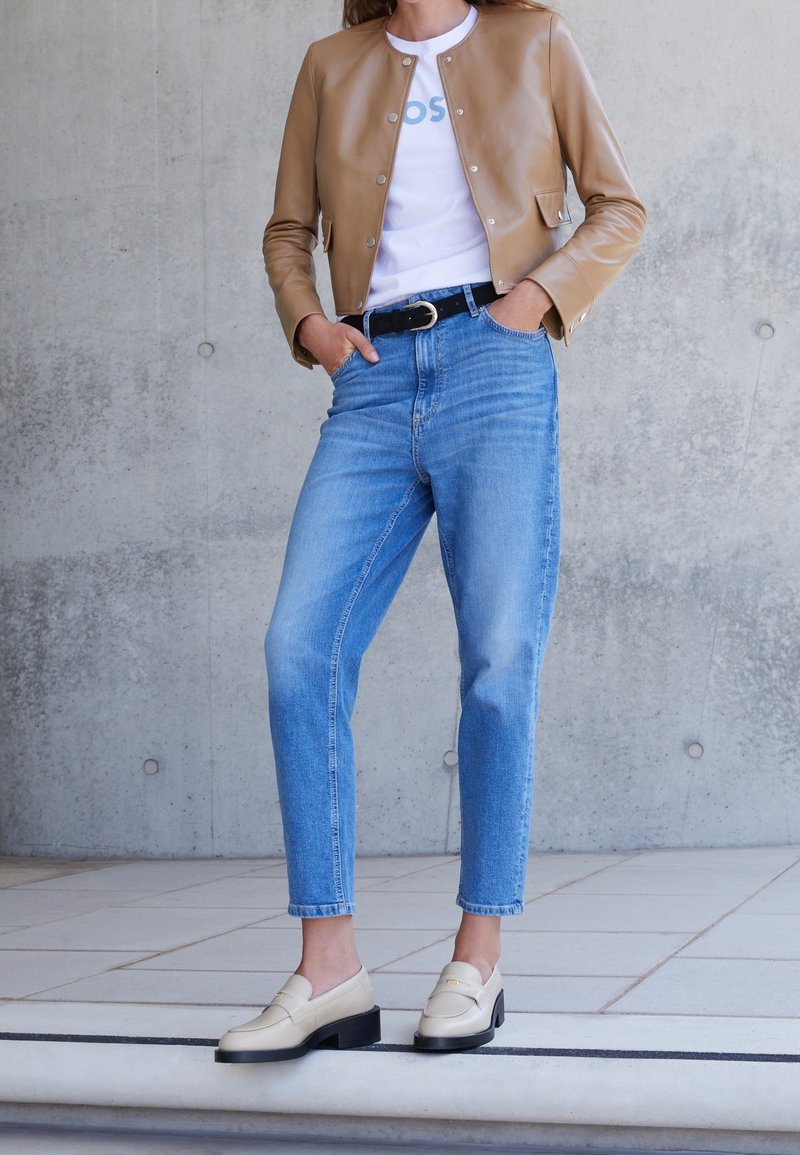 Person klädd i en ljusbrun kort läderjacka, vit t-shirt, högmidjade blå jeans, svart bälte och krämfärgade loafers som står på ett kaklat golv.