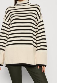 Przesadnie duży sweter w paski w kolorze kremowym i czarnym, z wysokim kołnierzem, prążkowaną teksturą i szerokimi rękawami. Pionowe paski przeplatają się różną grubością.