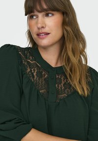 Tumši zaļa blūze ar mežģīņu apdari, noapaļotu apkakli un pogātām priekšpusi. Garas piedurknes ar savilktiem elementiem. Mīksta, teksturēta auduma.