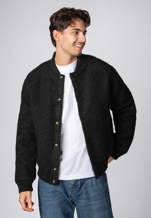 Schwarze, flauschige Bomberjacke mit geripptem Kragen und Bündchen. Mit Druckknöpfen und einem lockeren Schnitt, getragen über einem weißen T-Shirt und blauen Jeans.