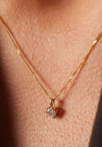 Collier en or avec un pendentif rond et transparent dans un sertissage à quatre griffes, reposant sur une peau avec une texture visible.