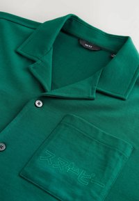 Chemise en coton verte avec un col cranté, une poche avant avec un texte discret, et deux boutons gris. Texture lisse avec un design décontracté.