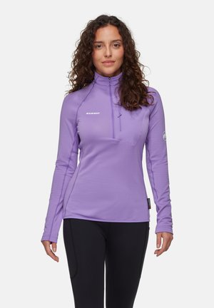 Maglietta sportiva a maniche lunghe di colore viola chiaro con zip a metà, design aderente, tessuto texturizzato e logo bianco sul petto. Pantaloni neri visibili.