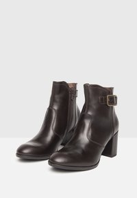 Botas de tobillo de cuero marrón con tacón ancho, cremallera lateral y un detalle de hebilla decorativa. Textura suave con un perfil elegante.