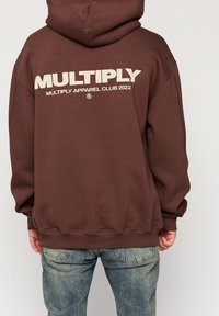 Felpa marrone scuro con un grande logo bianco "MULTIPLY" sul retro, dotata di un ampio cappuccio e polsini a coste, abbinata a jeans casual.