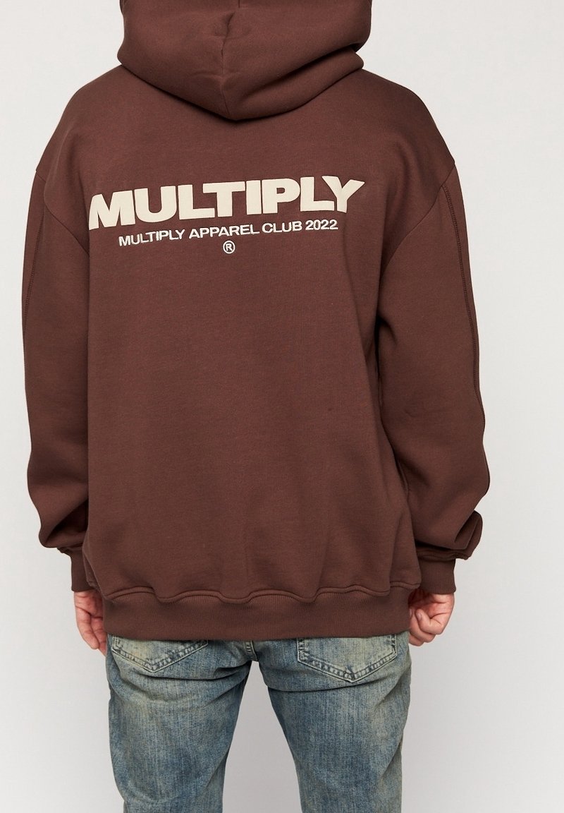 Felpa marrone scuro con un grande logo bianco "MULTIPLY" sul retro, dotata di un ampio cappuccio e polsini a coste, abbinata a jeans casual.