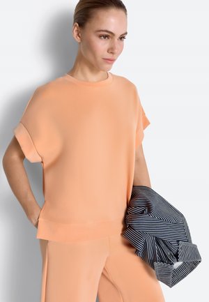 Ženska v breskovi kratkeročni majici in hlačah drži zloženo modro-belo črtasto majico na belem ozadju.