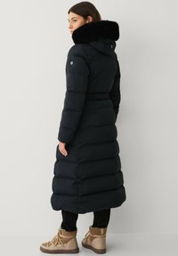 Zwarte lange pufferjas met een afneembare faux fur kraag, een getailleerde taille en een horizontaal gestikte ontwerp. De laarzen hebben veters.