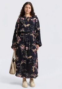Robe maxi fleurie à manches longues, fond bleu marine, taille élastique et motifs floraux colorés. Associée à des chaussures beige et un sac.