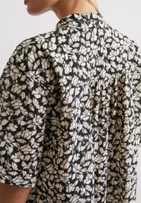 Blouse à motif floral noir et crème avec des manches courtes et un détail plissé dans le dos, en tissu léger.