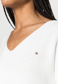 Witte gebreide trui met een V-halsontwerp, voorzien van een klein geborduurd logo in het blauw, rood en wit aan de linkerkant.