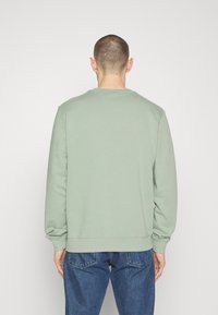 Sudadera verde clara con puños y cuello acanalados, de corte holgado y textura suave, usada sobre pantalones vaqueros azules.