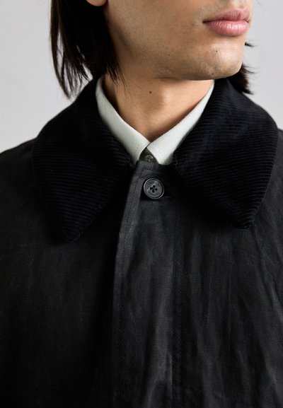 Collo in corduroy nero su una giacca scura, con chiusura a bottone e una camicia grigia chiara sotto; il tessuto ha una finitura opaca e testurizzata.