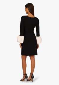 Czarna sukienka z długimi rękawami, z białymi, rozszerzanymi mankietami. Zamek błyskawiczny z tyłu. Model nosi czarne buty na wysokim obcasie. Gładka faktura.