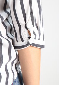 Blusa a righe bianche e grigie con un design delle maniche ampie, caratterizzata da un dettaglio a taglio al gomito. Tessuto morbido con una consistenza liscia.