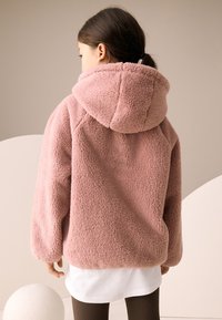 Vaaleanpunainen fleecetakki hupulla, jossa on pehmeä, teksturoitu pinta ja rento istuvuus, pidetty valkoisen paidan ja ruskeiden resoriboksihousujen päällä.