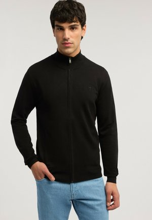 Junger Mann mit einem schwarzen, hochgeschlossenen Reißverschluss-Pullover und hellblauen Jeans, der mit einer Hand in der Tasche vor einem schlichten Hintergrund steht.