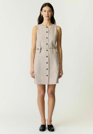 Kvinde stående med front mod kamera iført ærmeløs beige knap-ned kjole med bælte og sorte slip-on sko på hvid baggrund.