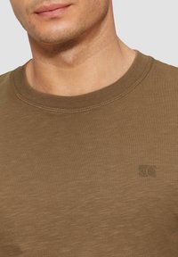 T-shirt olive green à col rond en tissu doux avec une texture lisse. Présente un petit logo sur la poitrine et un col côtelé.