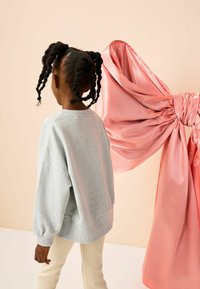 Felpa grigia con maniche larghe, abbinata a pantaloni beige a coste. Un grande pezzo di tessuto satinato rosa è appeso dietro, legato in un dettaglio a fiocco.