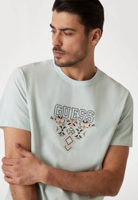 Světle modré tričko s logem "GUESS" a jemně vyšívanými květinovými motivy v růžové a černé barvě. Kulatý výstřih, krátké rukávy, měkká látka.