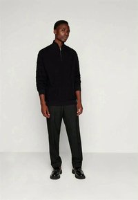 Black zip-up sweater with long sleeves, textured design. Paired with black trousers and black shoes on a neutral background.