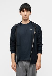 Μπλε σκούρο τζάκετ με φερμουάρ και υφή, σε συνδυασμό με ένα μονόχρωμο μπλε σκούρο μπλουζάκι. Γκρι παντελόνι ολοκληρώνει την εμφάνιση. Χαρακτηριστικά μινιμαλιστικού σχεδιασμού.