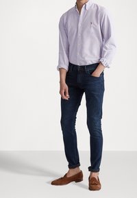 Muž v světle fialové košili na knoflíky, tmavě modrých slim-fit džínách a hnědých semišových mokasínech s třásněmi, stojící s jednou rukou v kapse.