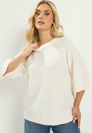 Blondynka ubrana w luźny, biały sweter dziany z rękawami do łokcia i niebieskie dżinsy, pozująca z jedną ręką przy dekolcie.