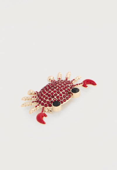 Broche en forme de crabe doré avec pierres précieuses rouges sur la carapace et pinces émaillées rouges, ornée de deux yeux en pierres précieuses noires, sur fond blanc.