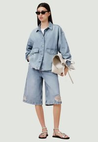 Sinine denimpusa, oversize-mudel, ja sobivad kulunud bermuda lühikesed püksid. Modell hoiab heledat kotti. Kanna musti päikeseprille ja rihmadega sandaale.