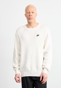 Valkoinen neulepaita, jossa on pitkät hihat ja ribbaustekniikalla valmistetut hihansuut. Vasemmassa rinnassa pieni musta Nike-logo. Yksinkertainen muotoilu, pehmeä rakenne.