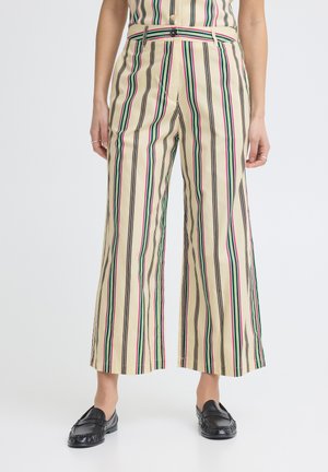 Pantaloni beige a gamba larga con strisce verticali nere, rosa e verdi, indossati con mocassini neri da una persona in piedi.