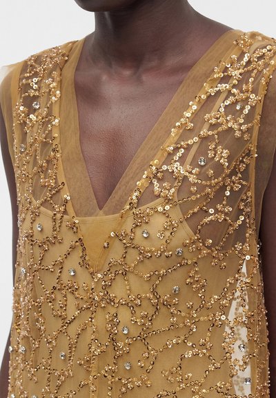 Gros plan sur une personne portant une robe dorée transparente ornée de broderies florales complexes en sequins et strass.