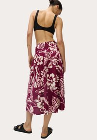 Gonna maxi bordeaux floreale con motivi tropicali bianchi, tessuto leggero, vita elastica, abbinata a un top corto nero e sandali a scivolo.