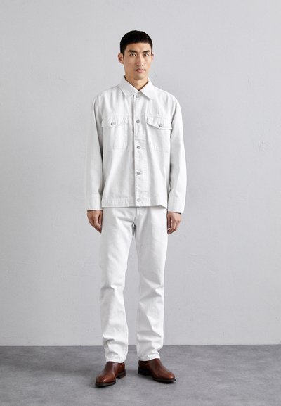 Veste en denim blanche avec deux poches poitrine, associée à un jean blanc. Des bottes en cuir marron complètent le look. Texture lisse, design minimaliste.