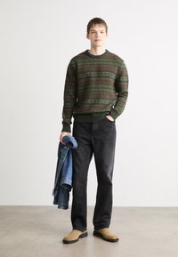 Knit sweater in dark green with multicolored geometric patterns, paired with black jeans and tan slip-on shoes, holding a denim jacket.