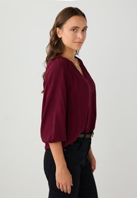 Frau mit langen braunen Haaren, die eine burgunderfarbene Bluse mit Puffärmeln und schwarze Hosen trägt, steht seitlich vor einem schlichten Hintergrund.