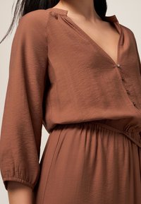 Blusa marrone leggera con scollo a V, dettaglio dei bottoni e maniche a tre quarti. Vita arricciata per una vestibilità rilassata. Tessuto morbido e testurizzato.