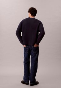 Maglione blu navy con collo tondo, abbinato a jeans blu scuro con due tasche posteriori. L'outfit è completato da scarpe nere.