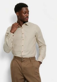 Hellbeige langärmeliges Hemd mit Kragen, durchgehender Knopfleiste und braunen Knöpfen. Kombiniert mit hellbraunen Hosen mit Bügelfalten.