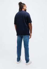 Άνδρας με dreadlocks που φοράει μπλε σκούρο T-shirt, μπλε τζιν και λευκά αθλητικά παπούτσια, στέκεται με την πλάτη προς τα εμπρός σε απλό λευκό φόντο.