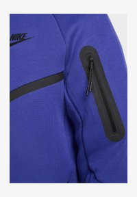 Lila Sweatshirt mit einem schwarzen Horizontalstreifen, ausgestattet mit einer Reißverschlusstasche am Ärmel und dem aufgedruckten Nike-Logo auf der Brust.