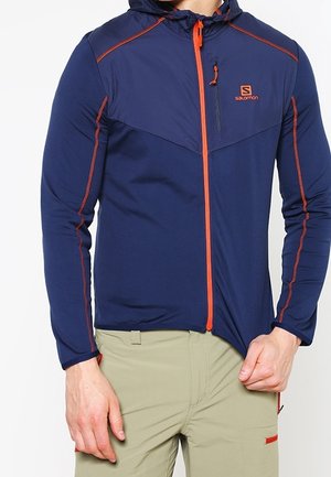 Man wearing navy blue Salomon jacket with orange zipper and stitching, paired with beige pants, pulling jacket hem down.