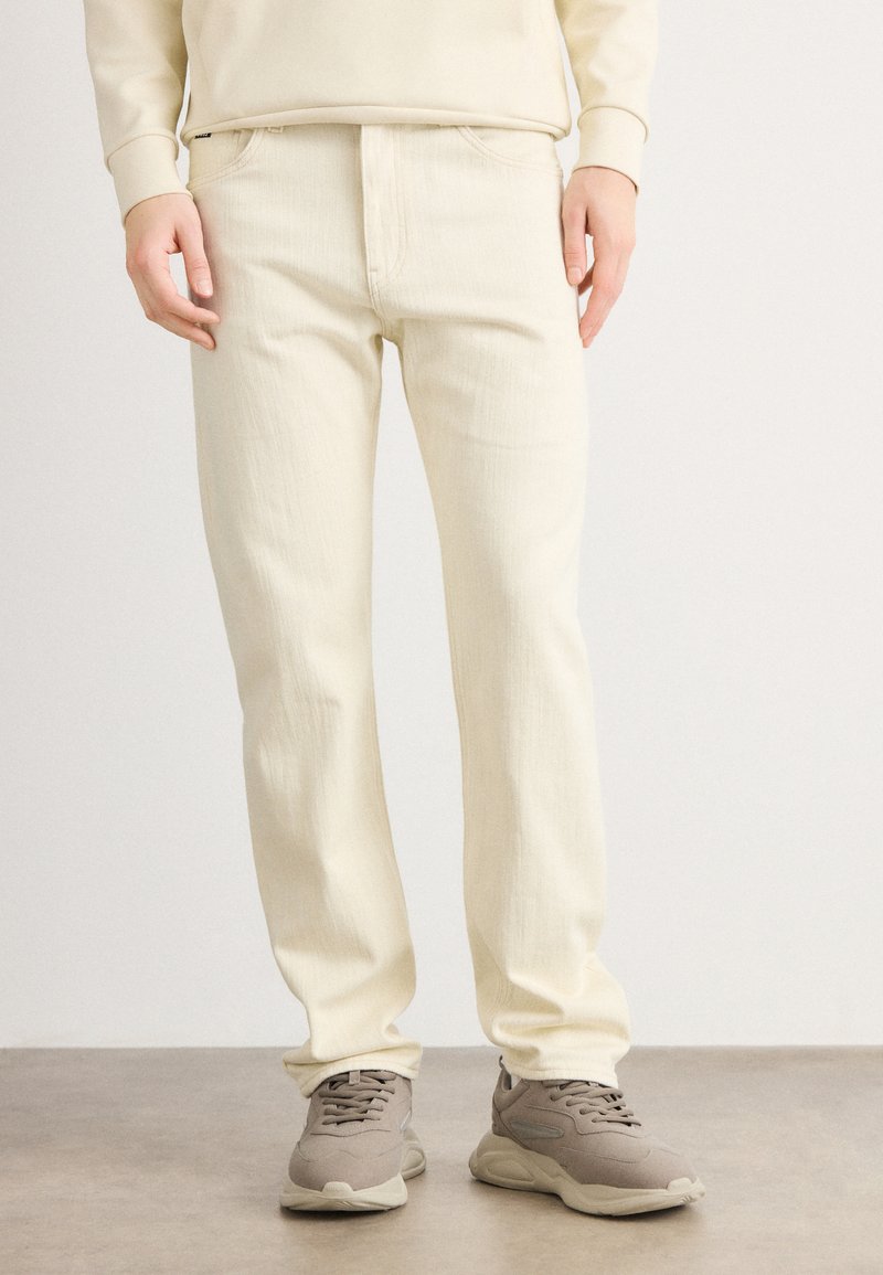 Homme portant un jean droit blanc cassé et des baskets chunky taupe, debout sur un sol gris clair avec un fond blanc.